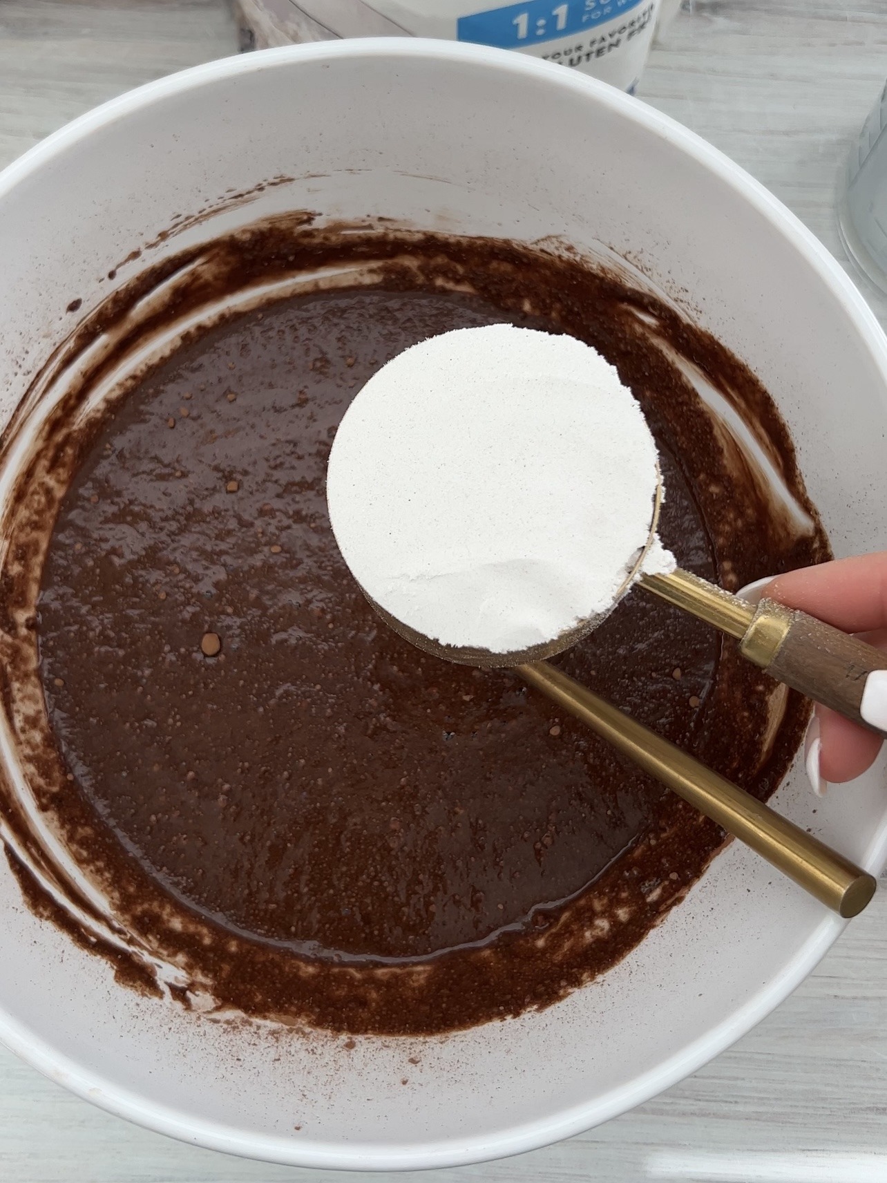 Chocolate cake batter in a white mixing bowl with a hand holding one cup of flour over top. 