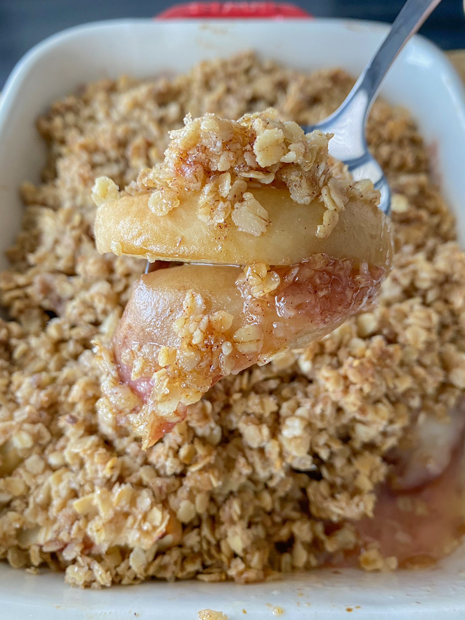 Silver fork with bite of peach crisp on it with the rest of the peach crisp in the background. 