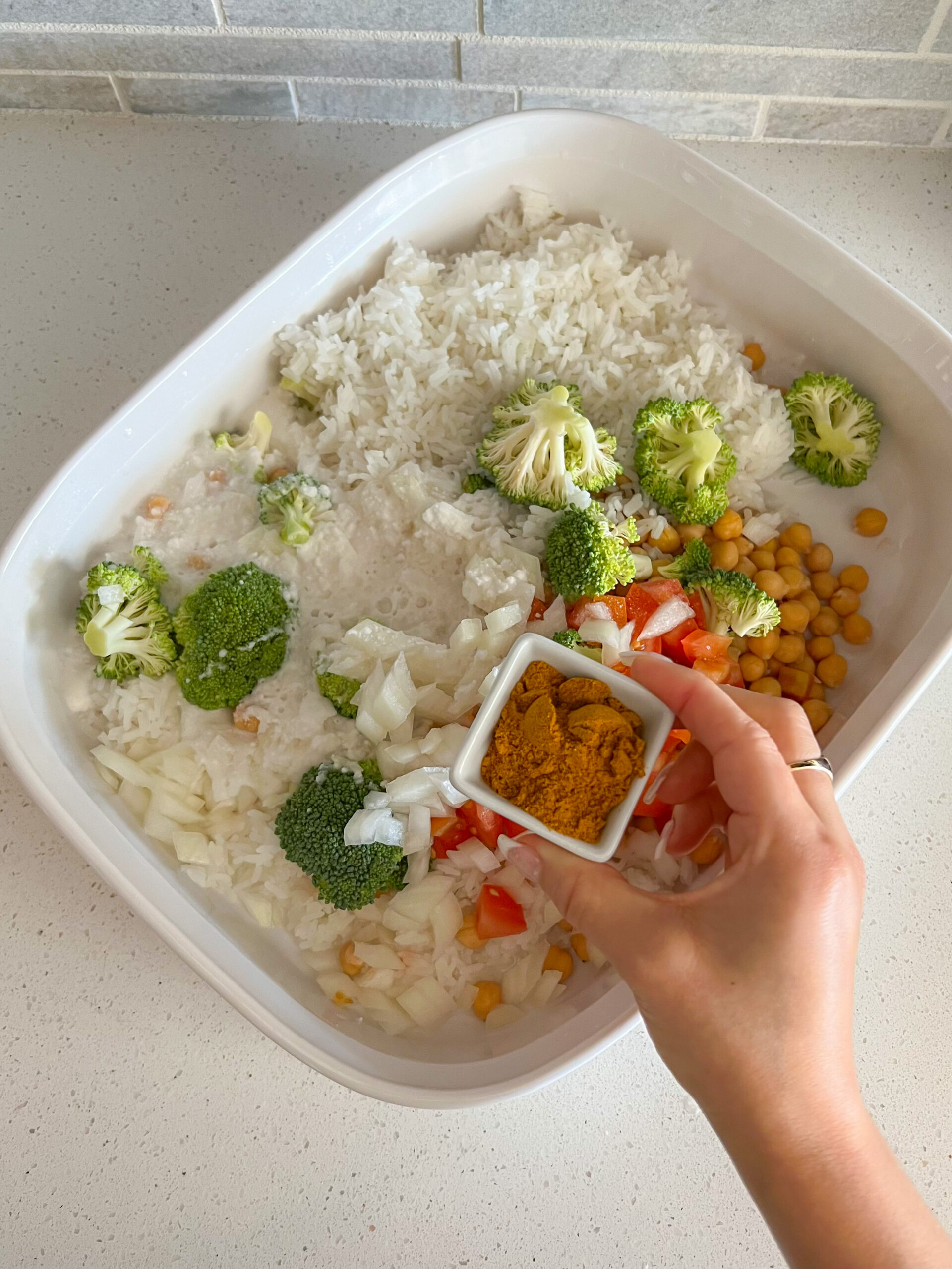 Rice and vegetable mixture in a casserole dish with a hand holding the spices above it.