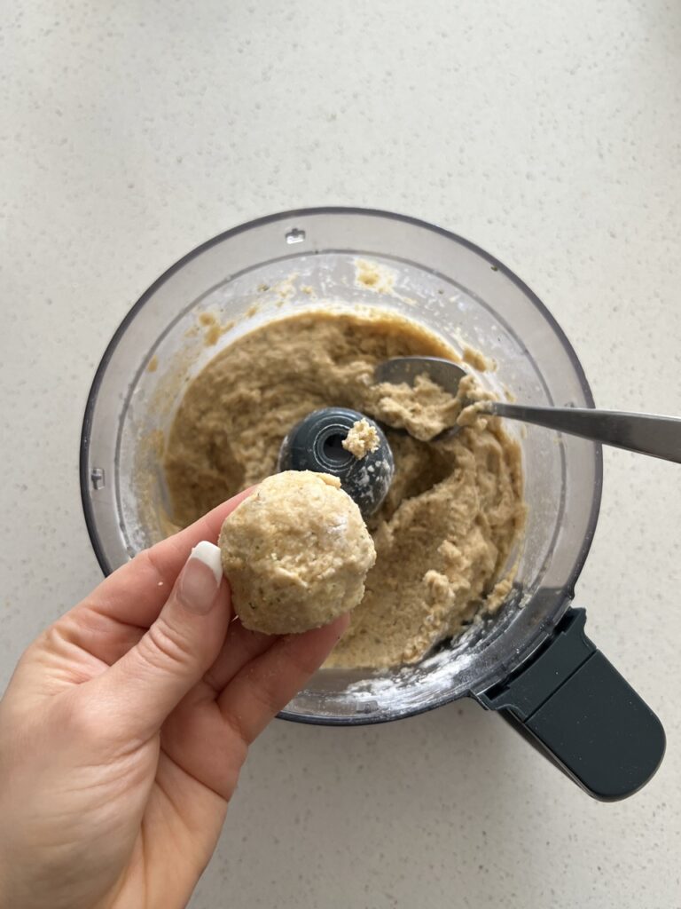 Chickpea meatball mixture in a food processor with a hand holding one meatball. 