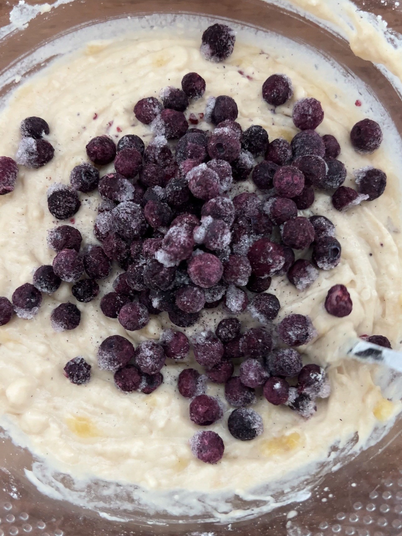 muffin batter in a clear mixing bowl with frozen blueberries being added.