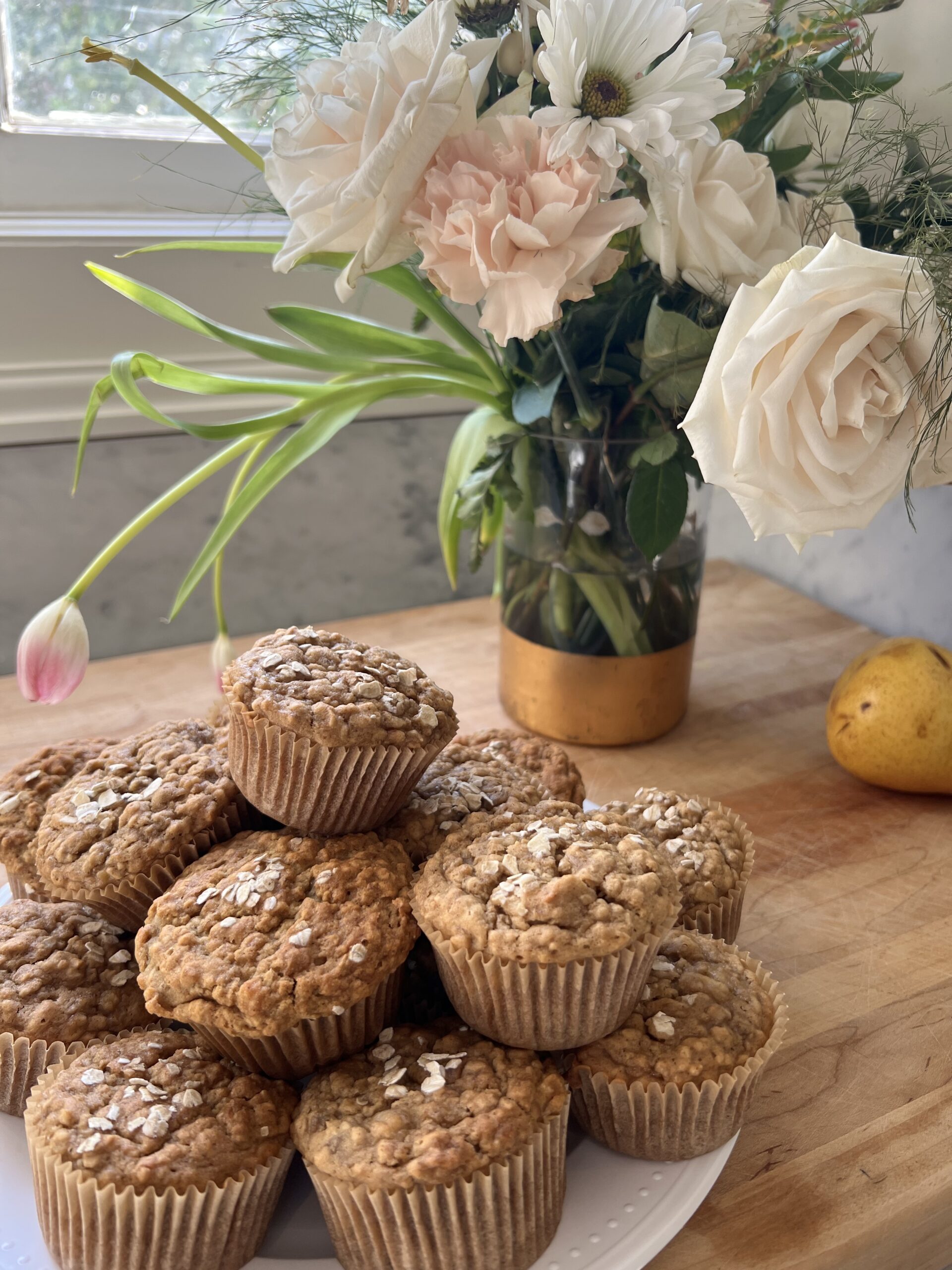 Banana muffins stacked on a white plate with a vase of flowers in the background. 